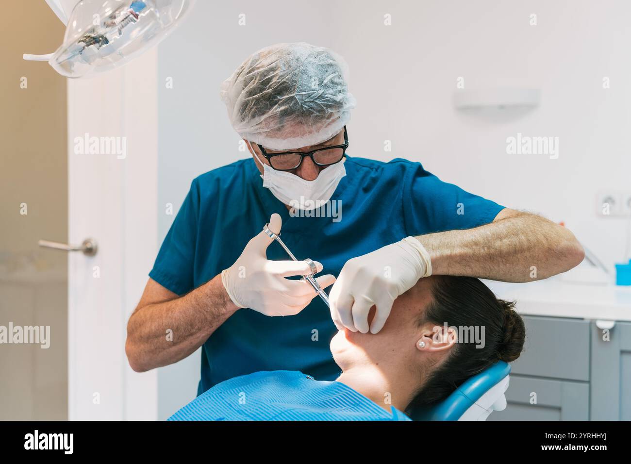 Zahnarzt in blauen Peelings und Maske bereitet sich auf die Narkose an einen Patienten vor. Die Umgebung ist eine moderne Zahnklinik mit einer hellen, sterilen Umgebung Stockfoto