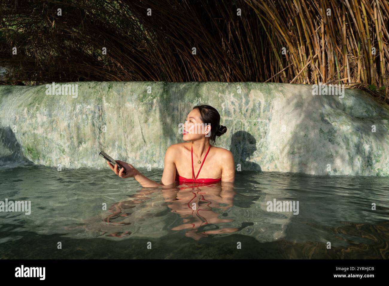 Eine asiatische Frau in rotem Badeanzug genießt ein friedliches Bad in der Terme di Saturnia, Toskana, Italien Sie hält ein Telefon, umgeben von natürlicher Schönheit und Ruhe Stockfoto