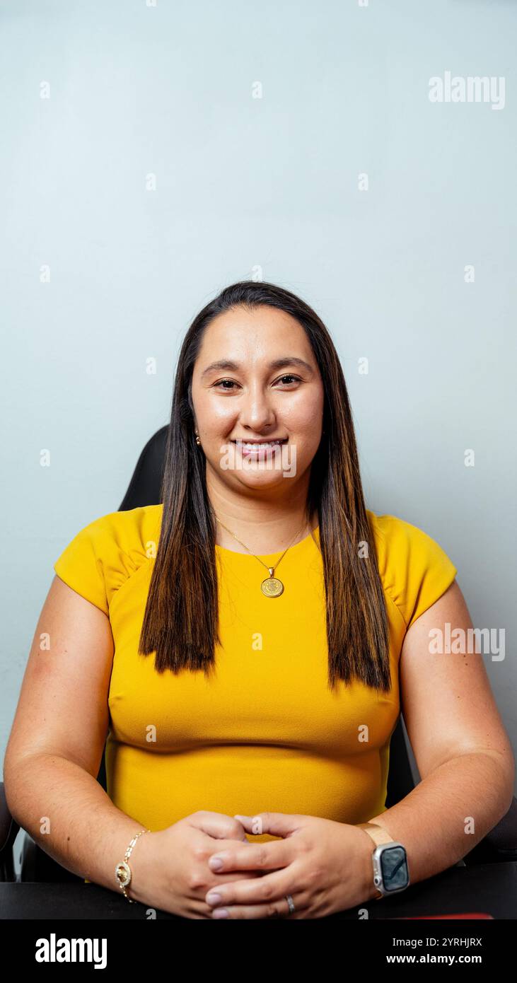 Eine selbstbewusste lateinfrau lächelt im Sitzen und trägt ein leuchtend gelbes Oberteil. Die Szene vermittelt ein professionelles Umfeld, das Positivität und App betont Stockfoto