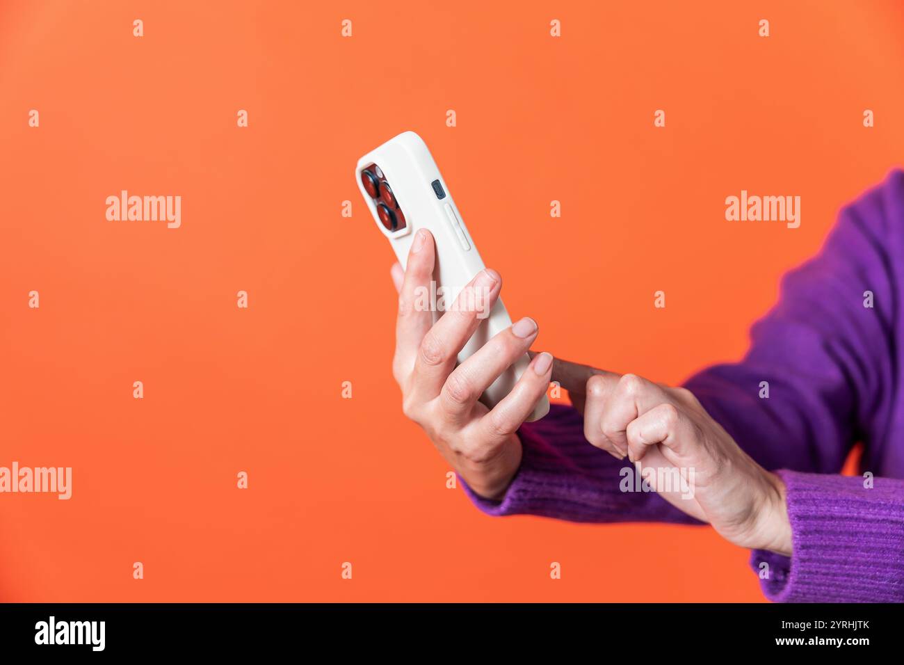 Dieses Bild zeigt eine ältere Erwachsene Frau in einem lila Pullover, die mit einem modernen Smartphone vor einem leuchtenden orangefarbenen Hintergrund interagiert. Der Fokus liegt auf h Stockfoto