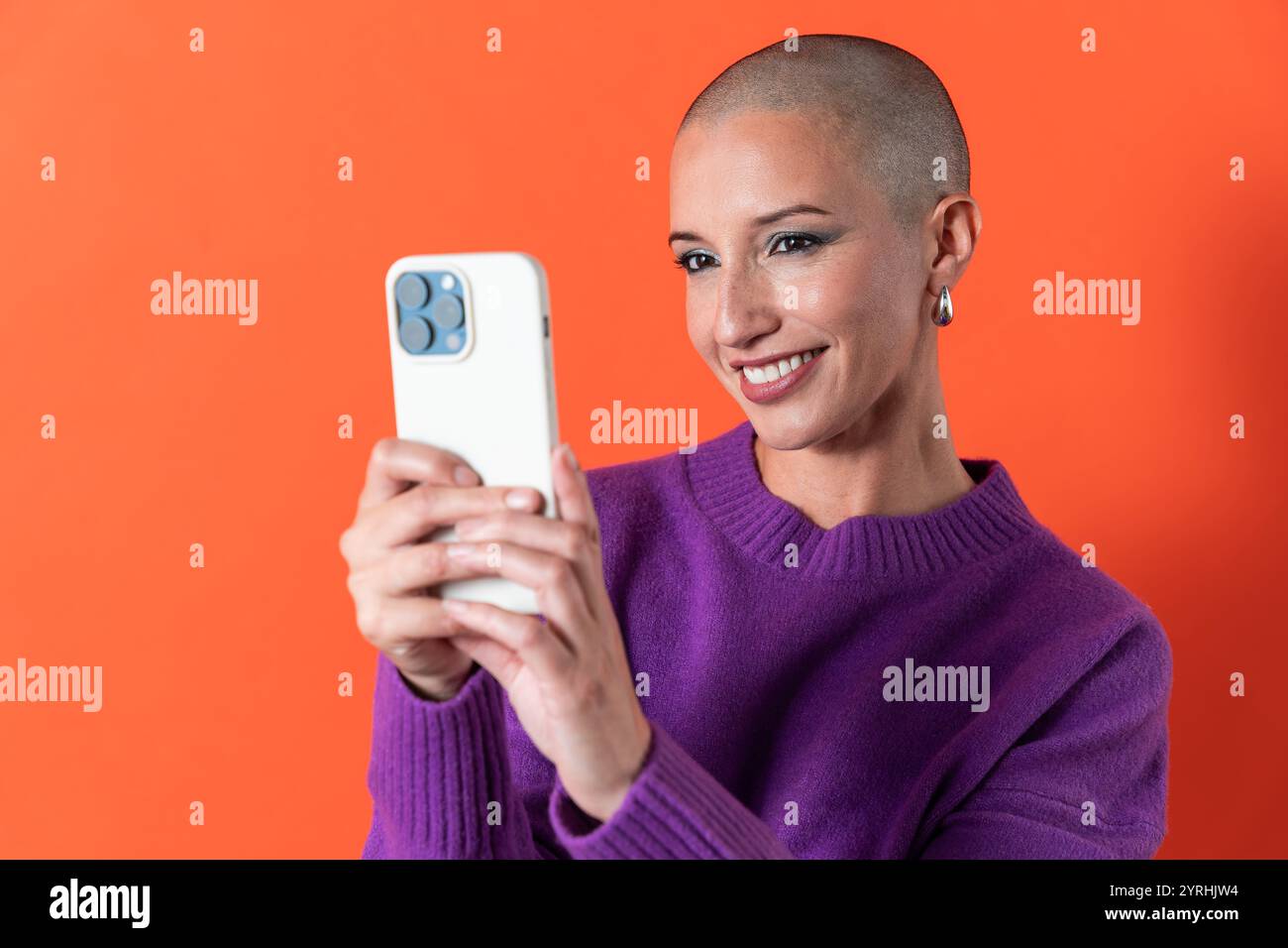 Eine selbstbewusste, kahlköpfige Erwachsene Frau in einem leuchtend violetten Pullover nutzt ihr Smartphone gerne vor einem leuchtend orangen Hintergrund. Ihre Freude zeigt sich in ihrem Lächeln Stockfoto