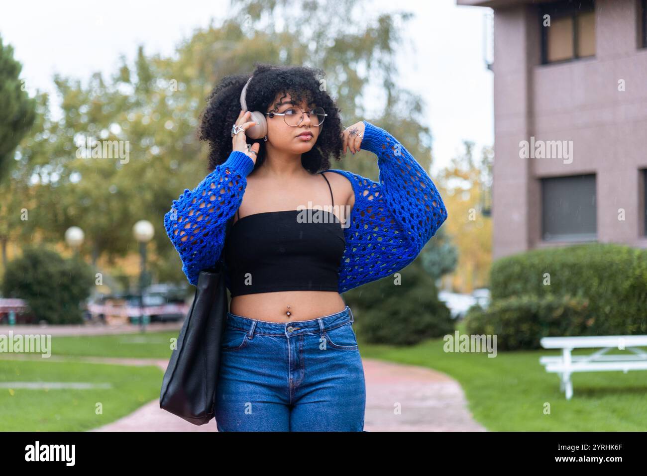 Afroamerikanerin mit Afro-Haaren, Kopfhörern, spaziert durch einen Universitätscampus Sie ist in lässiger Kleidung und zeigt eine lebendige und dynamische Atmosphäre Stockfoto