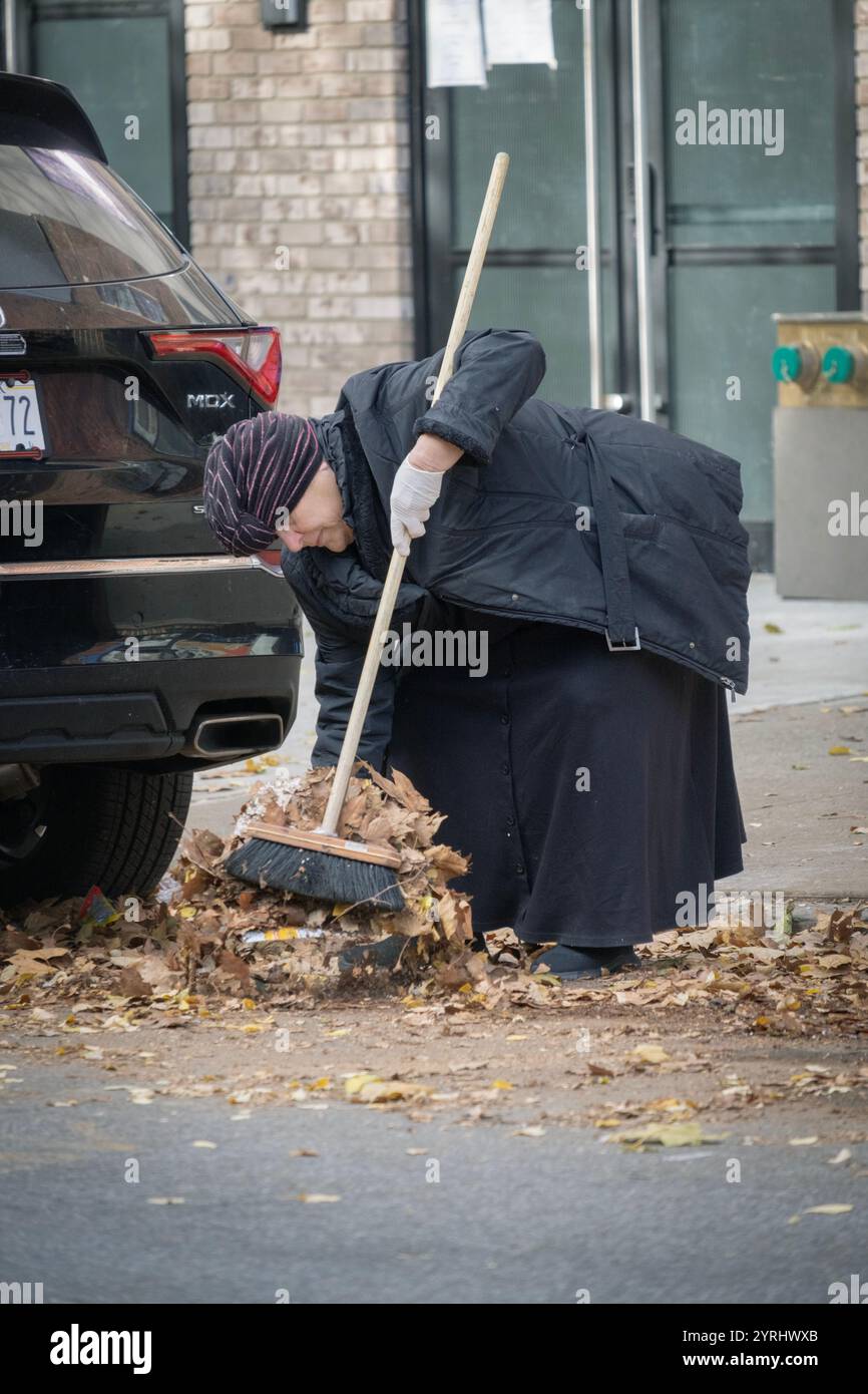 Eine bescheiden gekleidete orthodoxe Jüdin räumt Blätter vor ihrem Geschäft vom Gehweg. In einer Seitenstraße in Brooklyn, New York. Stockfoto