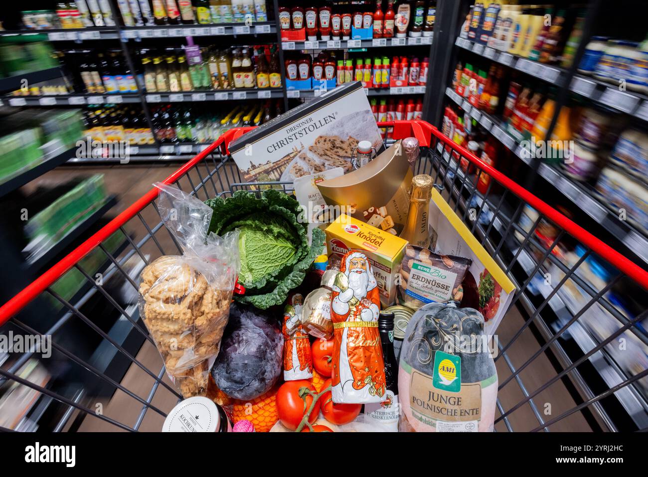 Köln, Deutschland. Dezember 2024. Ein vollständiger Warenkorb mit einer polnischen Ente, Gemüse, Obst, Weihnachtsstollen, Klößchen, Sekt, Wein, Pralinen und Schokoladen-Santas werden an Regalen in einem Supermarkt vorbei geschoben (verschwimmen aufgrund der langen Verschlusszeit). (Zur dpa-Umfrage: „Jede zweite Person spart Weihnachtseinkäufe“) Credit: Rolf Vennenbernd/dpa/Alamy Live News Stockfoto