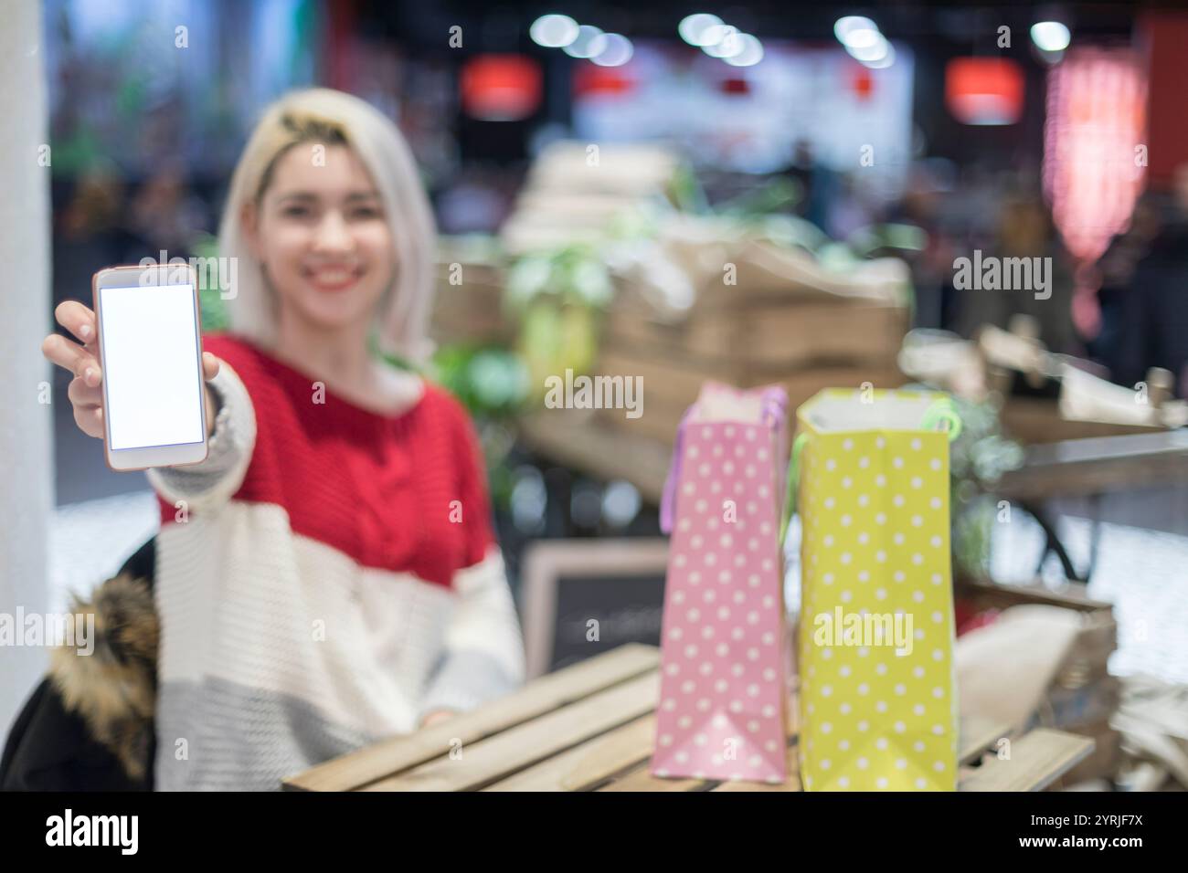 Eine lächelnde weiße Frau mit blonden Haaren präsentiert ihr Smartphone mit einem leeren Bildschirm. Sie sitzt drinnen mit Einkaufstaschen auf einem Holztisch nea Stockfoto