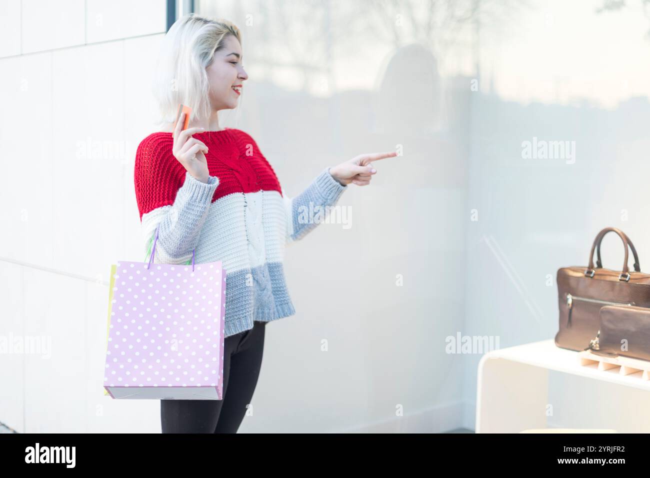 Eine junge Kaukasierin in einem roten Pullover zeigt auf eine Produktausstellung, während sie eine Einkaufstasche und eine Kreditkarte in der Hand hält, und zeigt einen modernen Einzelhandelsexperten Stockfoto