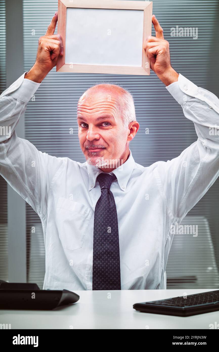 Reifer Geschäftsmann sitzt an seinem Schreibtisch, hält einen leeren Rahmen über dem Kopf, während er mit humorvollem Ausdruck auf die Kamera blickt und kreative Einladungen einlädt Stockfoto