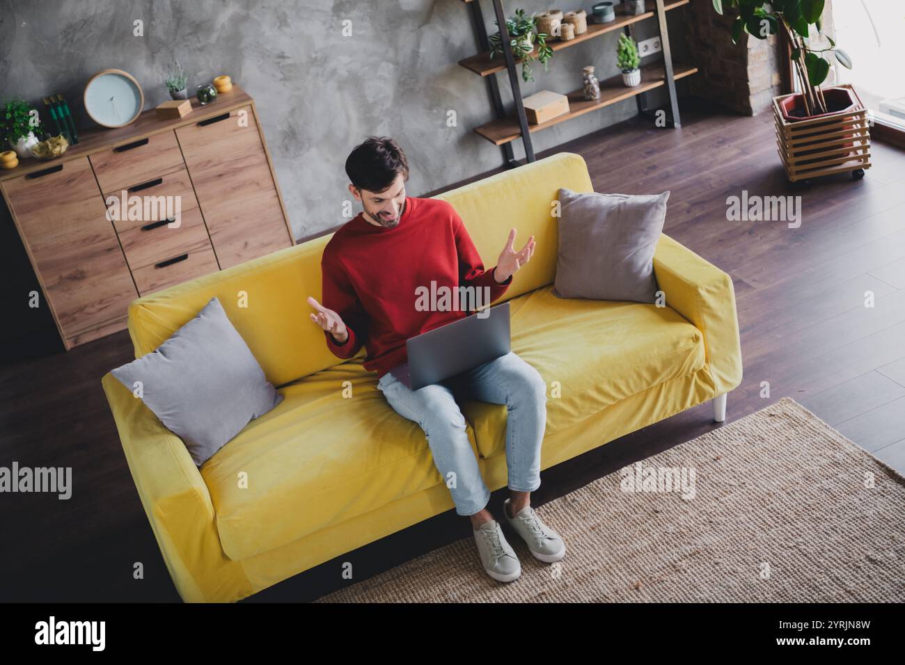 Junger Mann, der ein entspanntes Wochenende genießt, auf einem gelben Sofa sitzt, an einem Laptop in einer komfortablen Wohnung mit Tageslicht arbeitet. Stockfoto