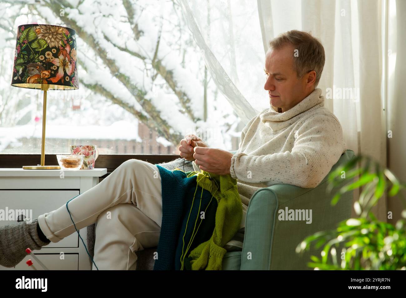 Mann im mittleren Alter sitzt zu Hause in einem Stuhl am Fenster, trägt einen warmen Pullover und strickt mit Nadeln. Gemütliches Zimmer mit schneebedecktem Winterblick Stockfoto