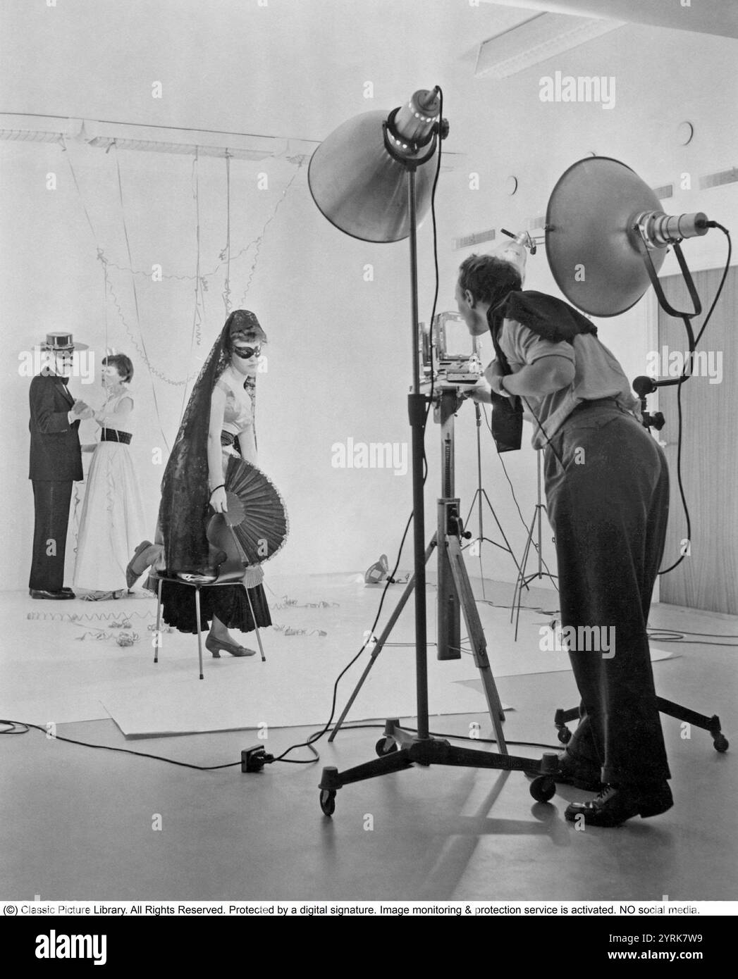 Fotograf und Models der 1950er Jahre. Eine junge Frau ist verkleidet, als wäre sie auf einer Maskerade-Party mit bedeckten Augen und spanischen Kleidern. Im Hintergrund tanzt ein Zorro-aussehender Mann mit einer Frau. Schweden 1957 Stockfoto