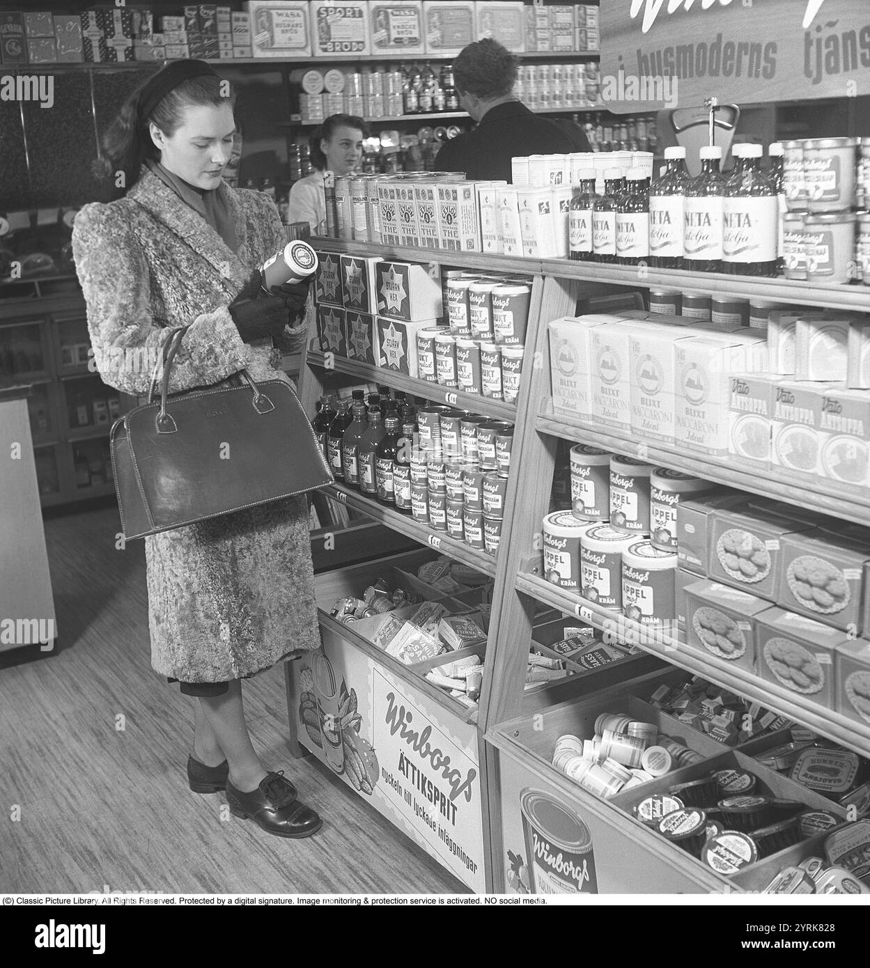 Eine Frau, die in den 1940ern Essen kauft Eine junge Frau kauft in einem Selbstbedienungsladen ein. Sie sieht sich eines von Winborgs Produkten genauer an, eine Obstmarmelade. Die Regale sind voller Pakete und Gläser und verschiedenen Speisen wie Makkaroni, Apfelsauce, Kochöl, Kekse usw. Schweden 1948. Kristoffersson Ref. AM81-2 Stockfoto