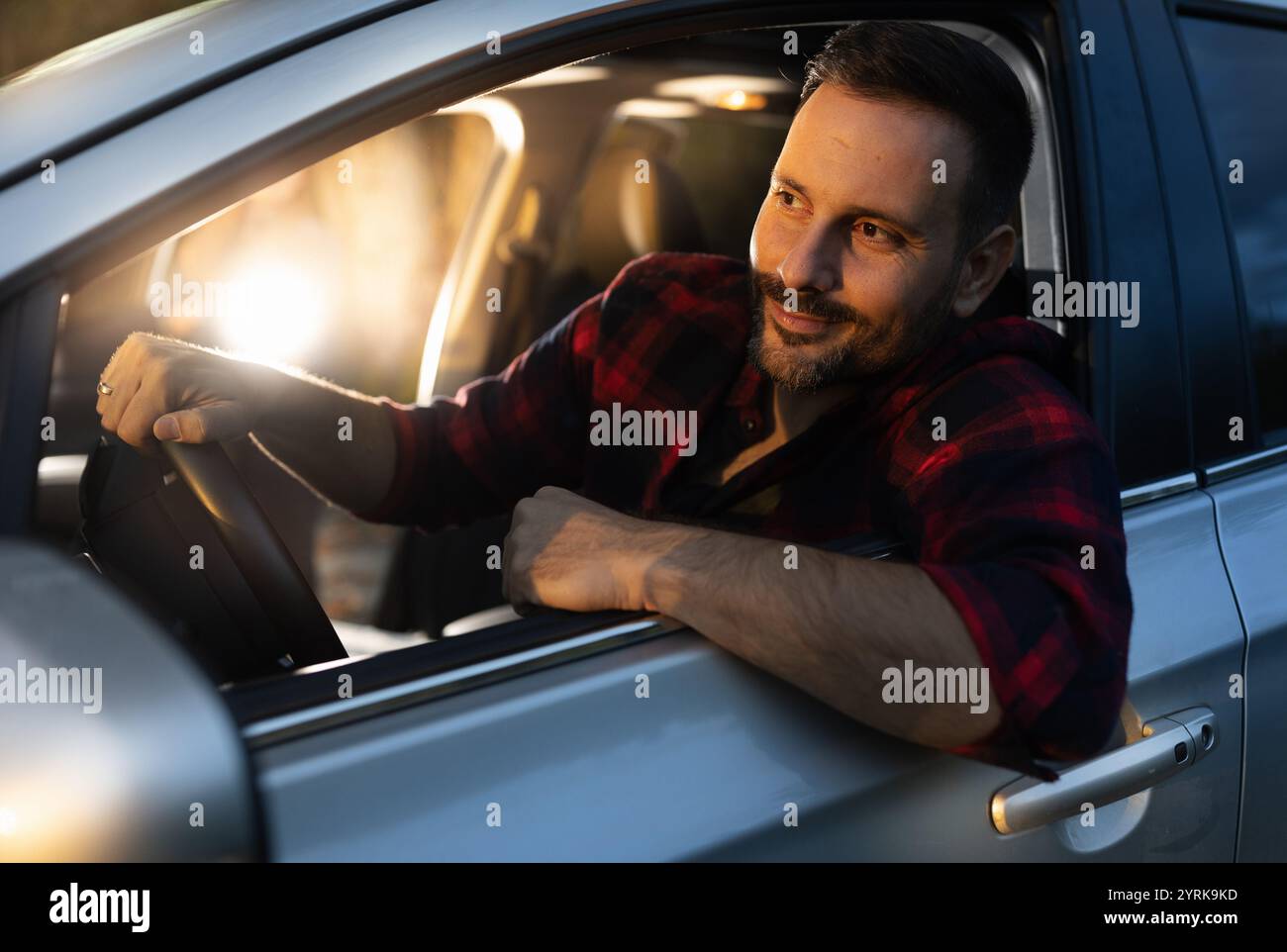 Fröhlicher Mann, der Auto fährt und durch Seitenfenster auf Sonnenuntergang auf der Landstraße nach vorne blickt Stockfoto