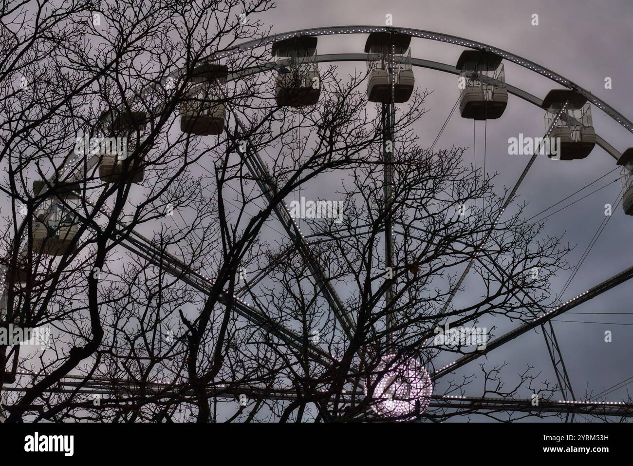 Der Bogen eines Riesenrads mit beleuchteten Gondeln wird teilweise von nackten Winterbäumen vor einem gedämpften Himmel verdeckt. Das Bild zeigt einen Kontrast zwischen dem Bild Stockfoto