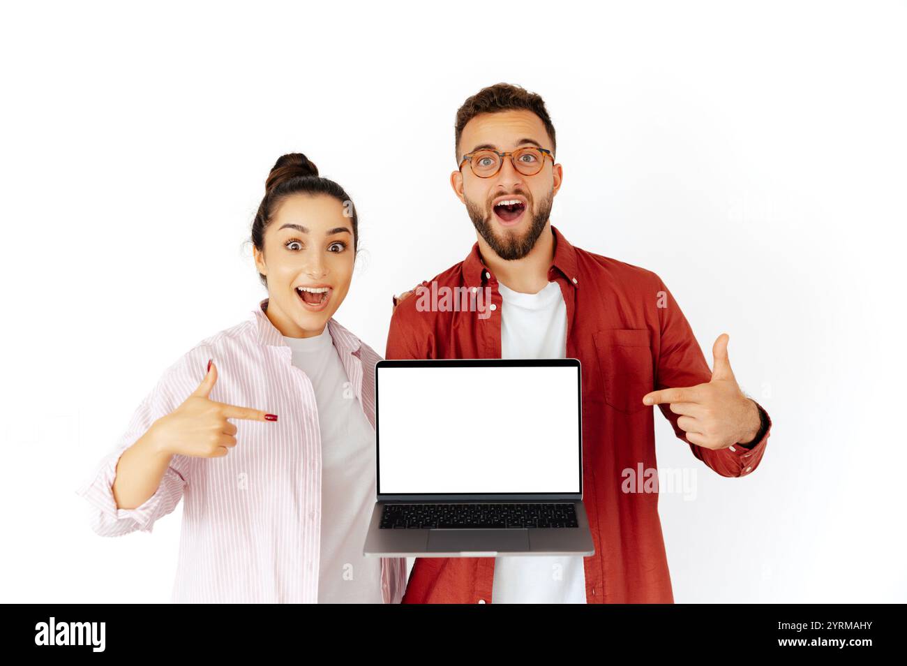 Aufgeregter Mann und Frau arabischer oder kaukasischer Herkunft, die einen Laptop mit leerem Modellbildschirm halten, lächeln und darauf zeigen, Inhalte oder Produkte bewerben, stehen auf einem isolierten weißen Hintergrund Stockfoto