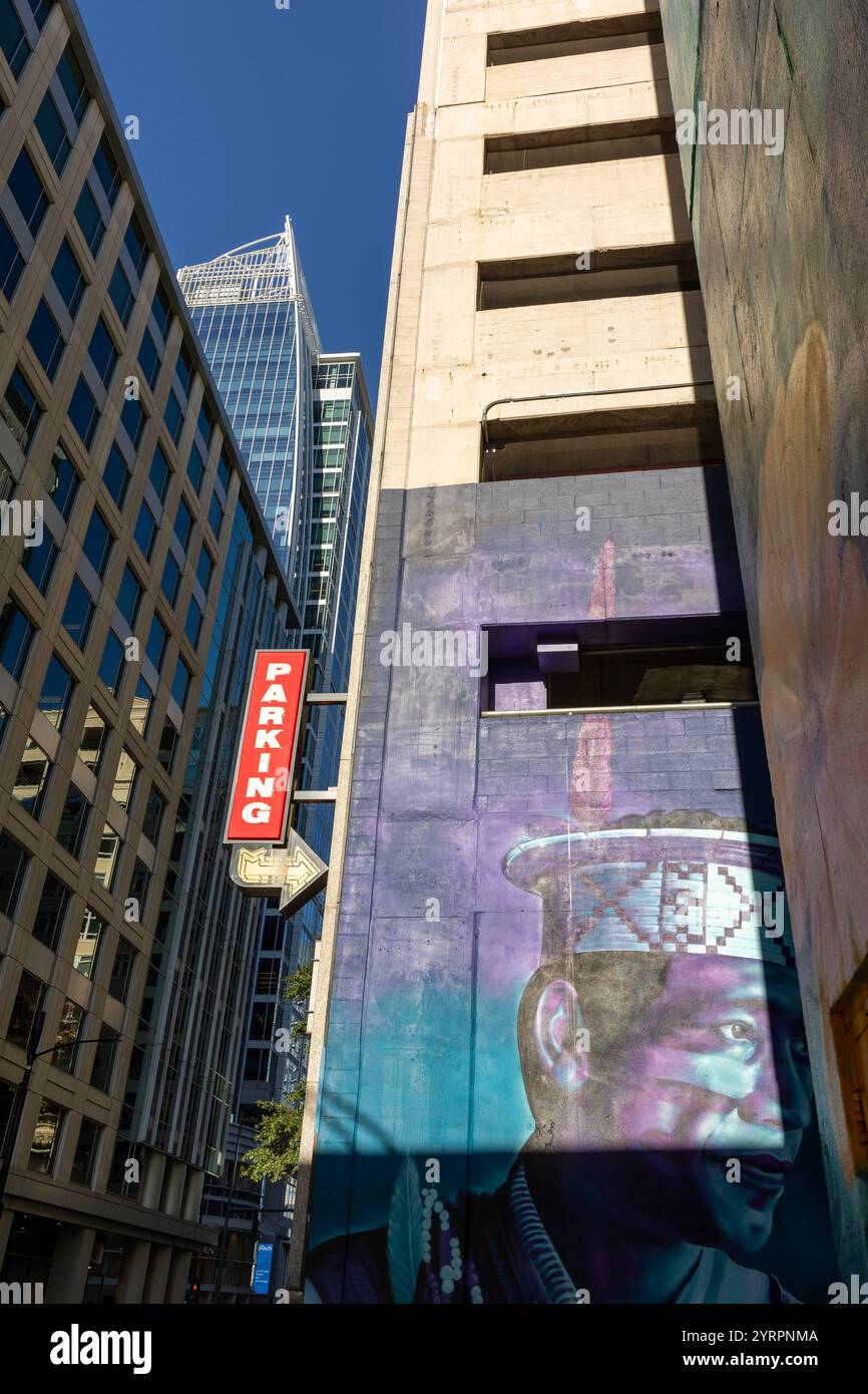 Eine beeindruckende Street-Art-Wandmalerei, die einen indigenen Mann mit traditionellem Kopfschmuck zeigt, schmückt die Fassade eines Parkhauses in Charlotte, North Carolina. Im Hintergrund ragen moderne Glas- und Betongebäude in den blauen Himmel, was einen faszinierenden Kontrast zwischen traditioneller Kunst und zeitgenössischer Architektur schafft. Das rote Parking-Schild verstärkt die urbane Atmosphäre der Szene. *** Ein beeindruckendes Street Art Wandgemälde, das einen Ureinwohner mit traditionellem Kopfschmuck zeigt, ziert die Fassade eines Parkhauses in Charlotte, North Carolina im Hinterland Stockfoto