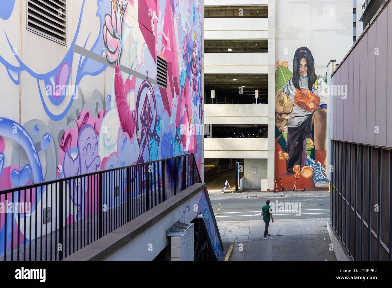 Das Foto zeigt eine beeindruckende Szene urbaner Kunst in Charlotte, North Carolina. Die Wände des schmalen Durchgangs sind mit farbenfrohen graffitis und großformatigen murals bedeckt, die eine dynamische und kreative Atmosphäre schaffen. Auf der linken Seite befinden sich verspielte, abstrakte Muster in leuchtenden Farben wie Pink, Blau und Lila. Die rechte Wand zeigt ein realistisches Wandbild mit einer jungen Frau, die eine Tasche mit Orangen trägt, in einer Szene voller Bewegung und Energie. Im Hintergrund ist ein mehrstöckiges Parkhaus sichtbar, das einen Kontrast zur lebendigen Straße A Stockfoto