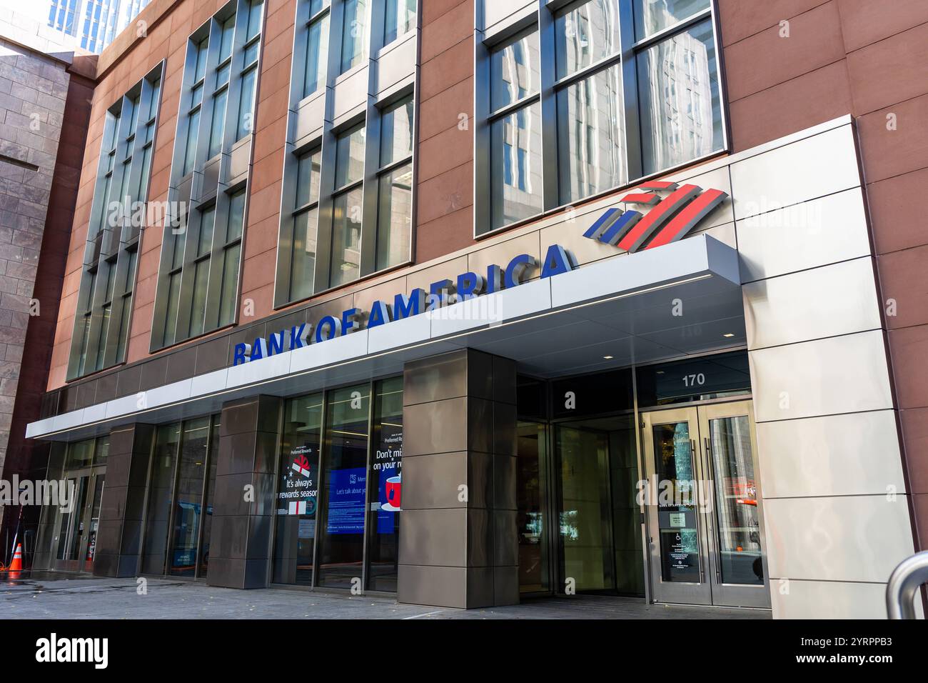 Das Foto zeigt den Eingangsbereich einer Bank of America Filiale in Charlotte, North Carolina. Die Fassade des Gebäudes ist modern gestaltet, mit großen Glasfenstern, die Umgebung reflektieren, sowie eine Kombination aus braunem und metallischem Material. Der Schriftzug Bank of America in blauen Buchstaben ist prominent über dem Eingang angebracht, begleitet vom ikonischen rot-blau-weißen Logo. Der Eingang selbst ist durch eine Glas-Doppeltür gekennzeichnet, auf der wichtige Informationen für Kunden sichtbar sind. Auf den Fenstern im Inneren sind Anzeigen mit Finanzinformationen zu sehen. Stockfoto