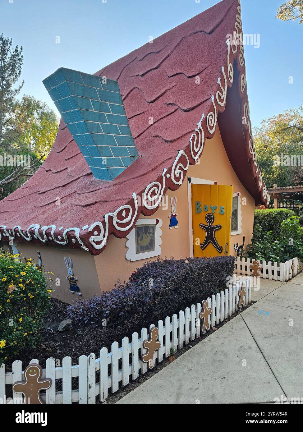 Ein skurriles Lebkuchenhaus mit Süßigkeiten-inspirierten Dekorationen, umgeben von einem weißen Streifenzaun mit Lebkuchenfiguren. - Smartphone-aufgenommenes Stockfoto