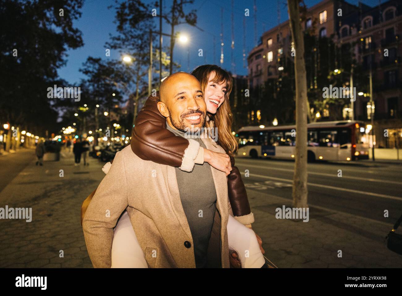 Ein lächelndes Paar erfreuen sich bei Nacht an einem Huckepack-Ritt entlang einer lebhaften Stadtstraße, um die romantische Abendatmosphäre zu verbessern. Stockfoto