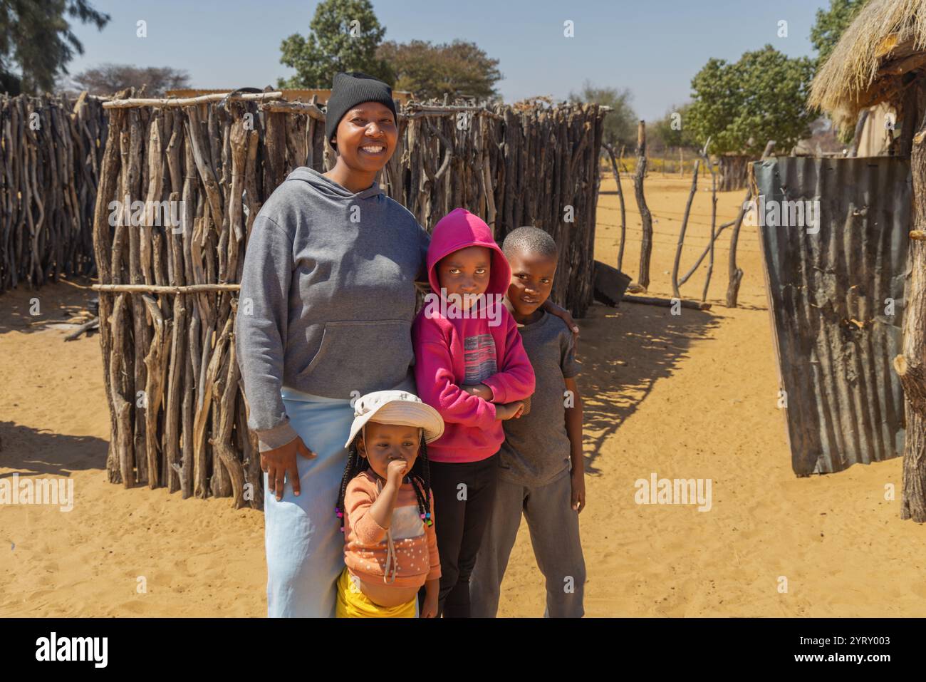 Dorf, Porträt afrikanischer Mutter mit drei Kindern, draußen auf dem Hof, Hütte und Holzzaun im Hintergrund, Stockfoto