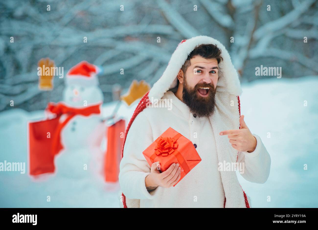 Weihnachten Winter Menschen Porträt. Mann mit rotem Geschenk, der mit Schneemann im Winterpark spielt. Schöner Weihnachtsmann. Schneemann und lustiger bärtiger Mann in der Stockfoto