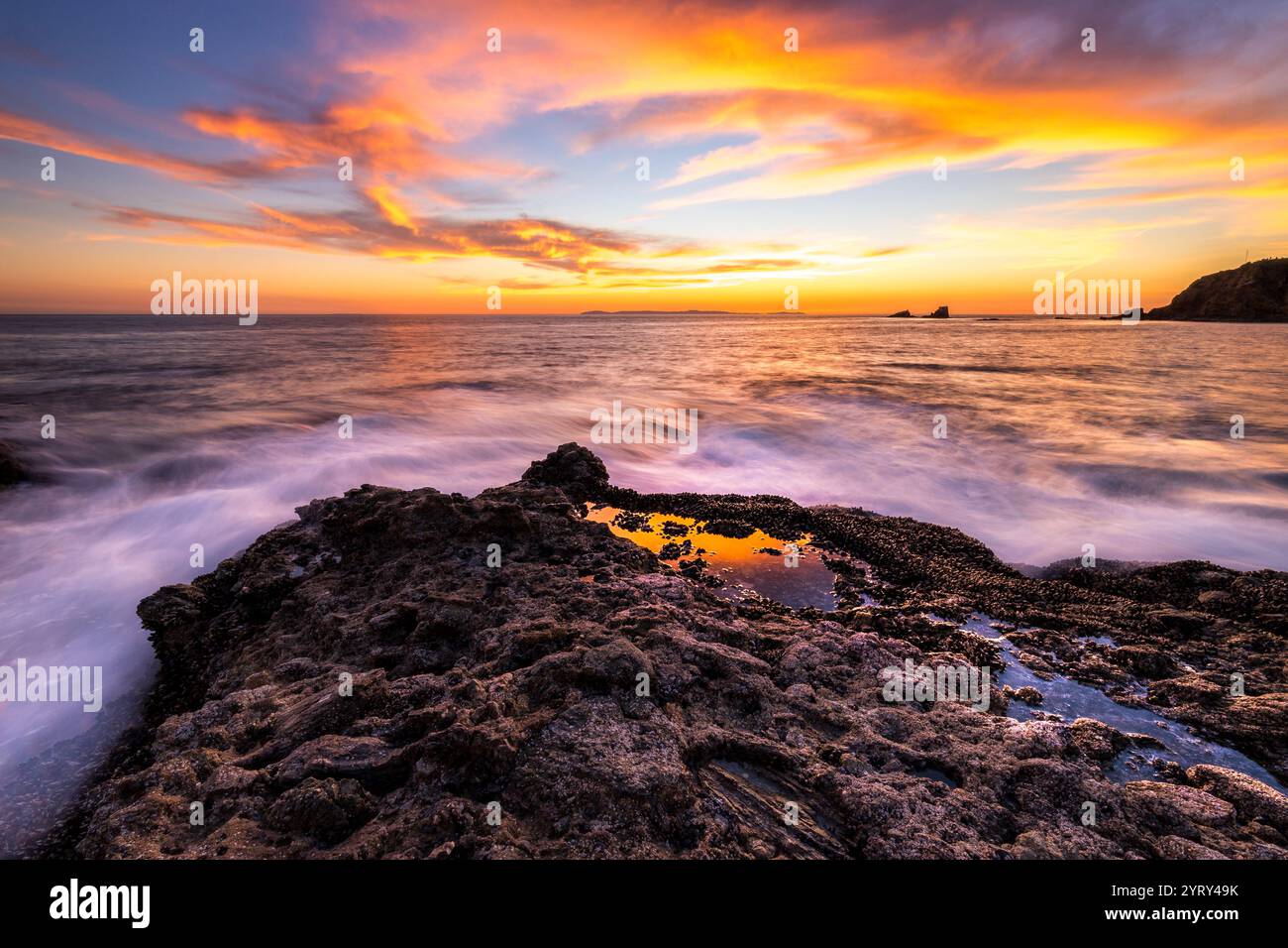 Wellen schlagen sanft gegen die felsige Küste am Crescent Bay Beach während eines pulsierenden Sonnenuntergangs in Laguna Beach. Der Himmel ist in warmen Farben gemalt. Stockfoto