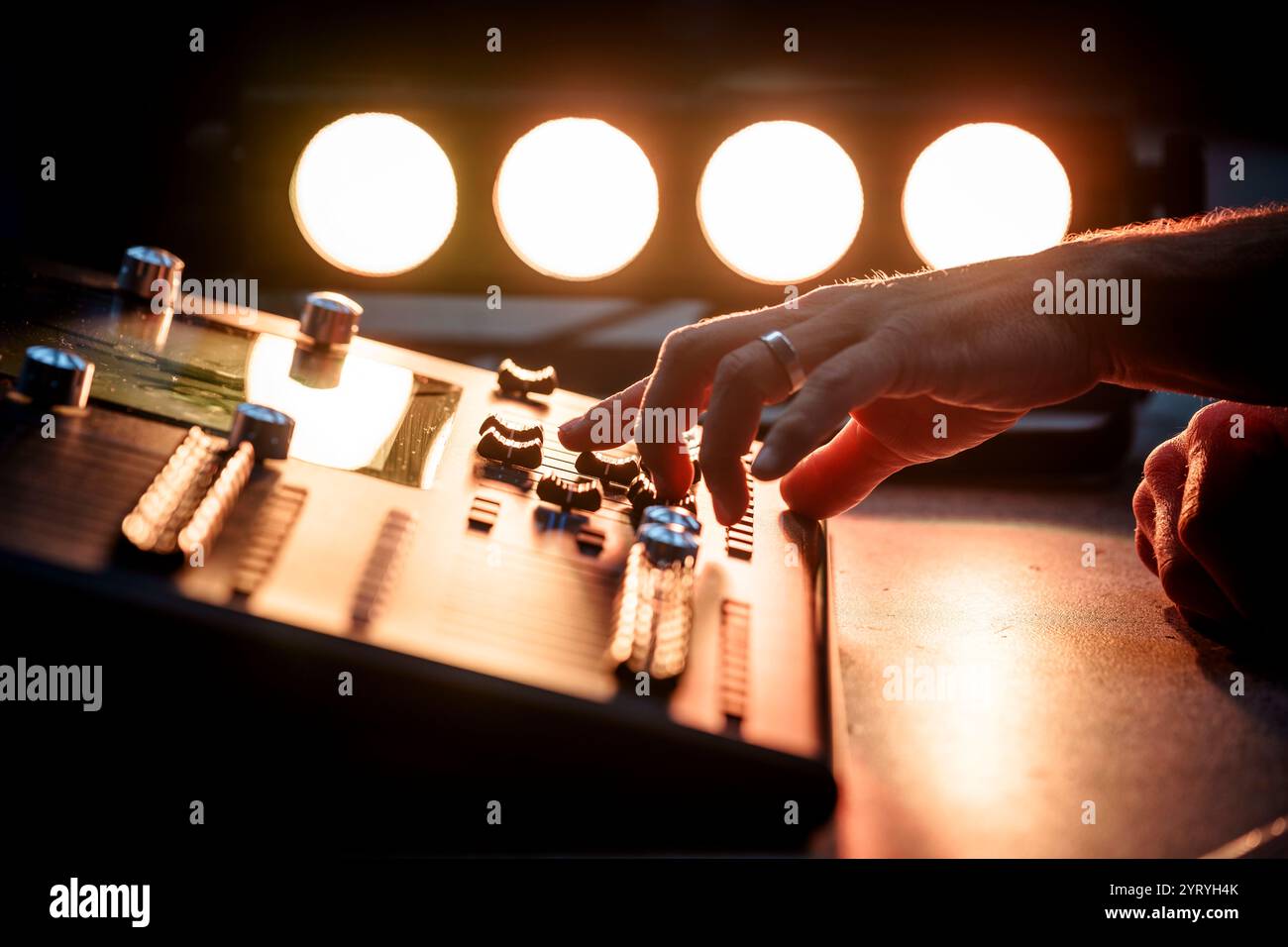 Nahaufnahme der Hände eines Technikers, der eine Tonmischkonsole bedient, die von Bühnenbeleuchtung beleuchtet wird und die Audioproduktion bei einer Live-Veranstaltung betont. Stockfoto