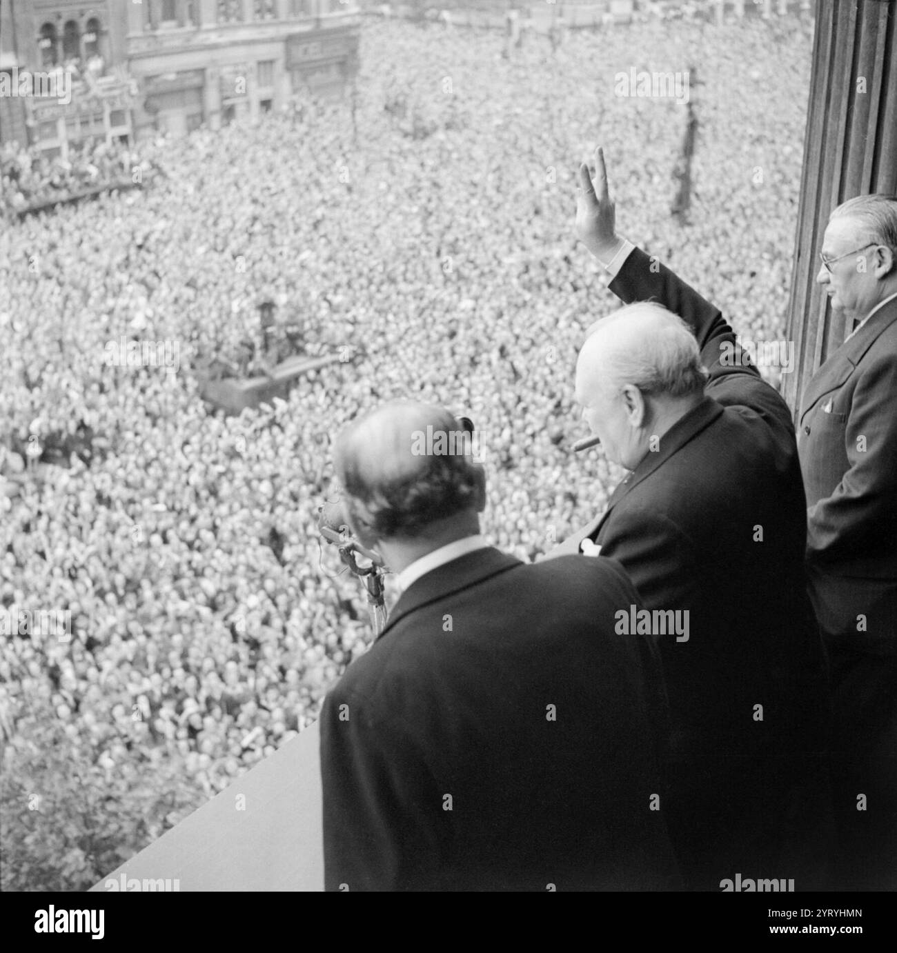 Winston Churchill winkt den Menschenmassen in Whitehall in London zu, während sie den VE Day am 8. Mai 1945 feiern. Vom Balkon des Gesundheitsministeriums überreicht Premierminister Winston Churchill den Menschenmassen in Whitehall sein berühmtes „V for Victory“-Schild an dem Tag, an dem er der Nation den Sieg des Krieges mit Deutschland übermittelte, am 8. Mai 1945 (VE Day). Zu Churchills Linker gehört Sir John Anderson, der Finanzminister. Zu Churchills Rechten ist Ernest Bevin, der Arbeitsminister. Stockfoto