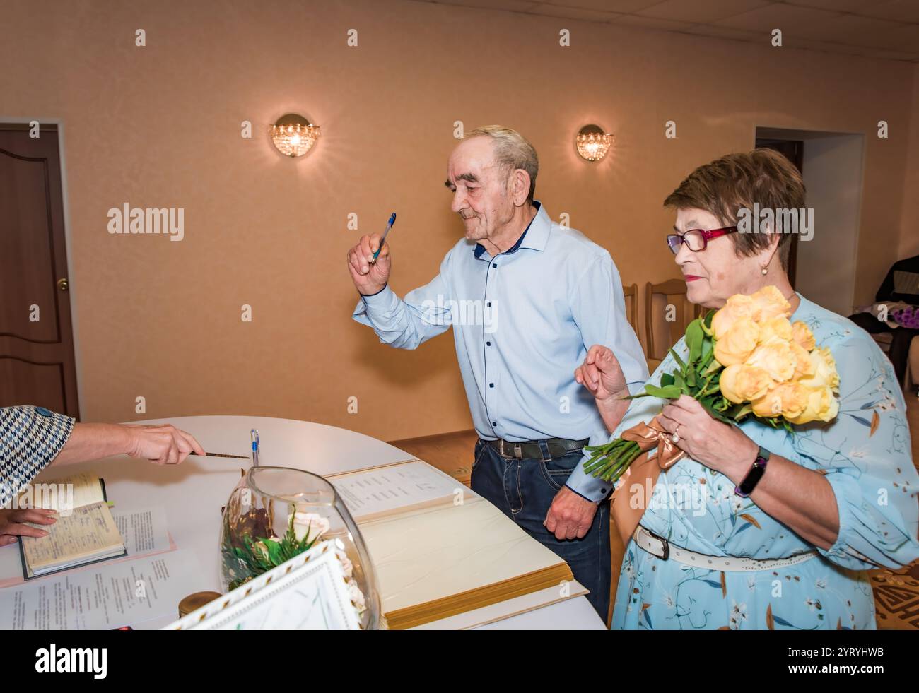 Ältere russische Paare in ihren 70ern feiern ihren 50. Hochzeitstag, indem sie Dokumente in einem Standesamt unterschreiben. Die Frau hält einen Strauß davon Stockfoto