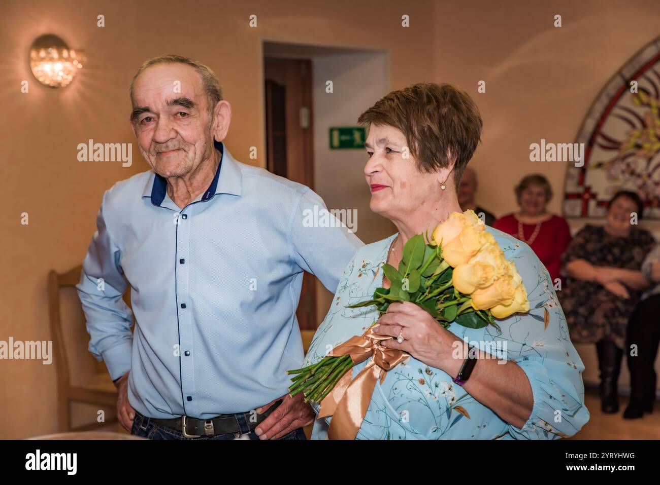 Russische ältere Ehepaare, beide in ihren 70ern, feiern ihre Hochzeit in einem Standesamt. Die Braut hält einen Strauß gelber Rosen, während Freunde sitzen Stockfoto