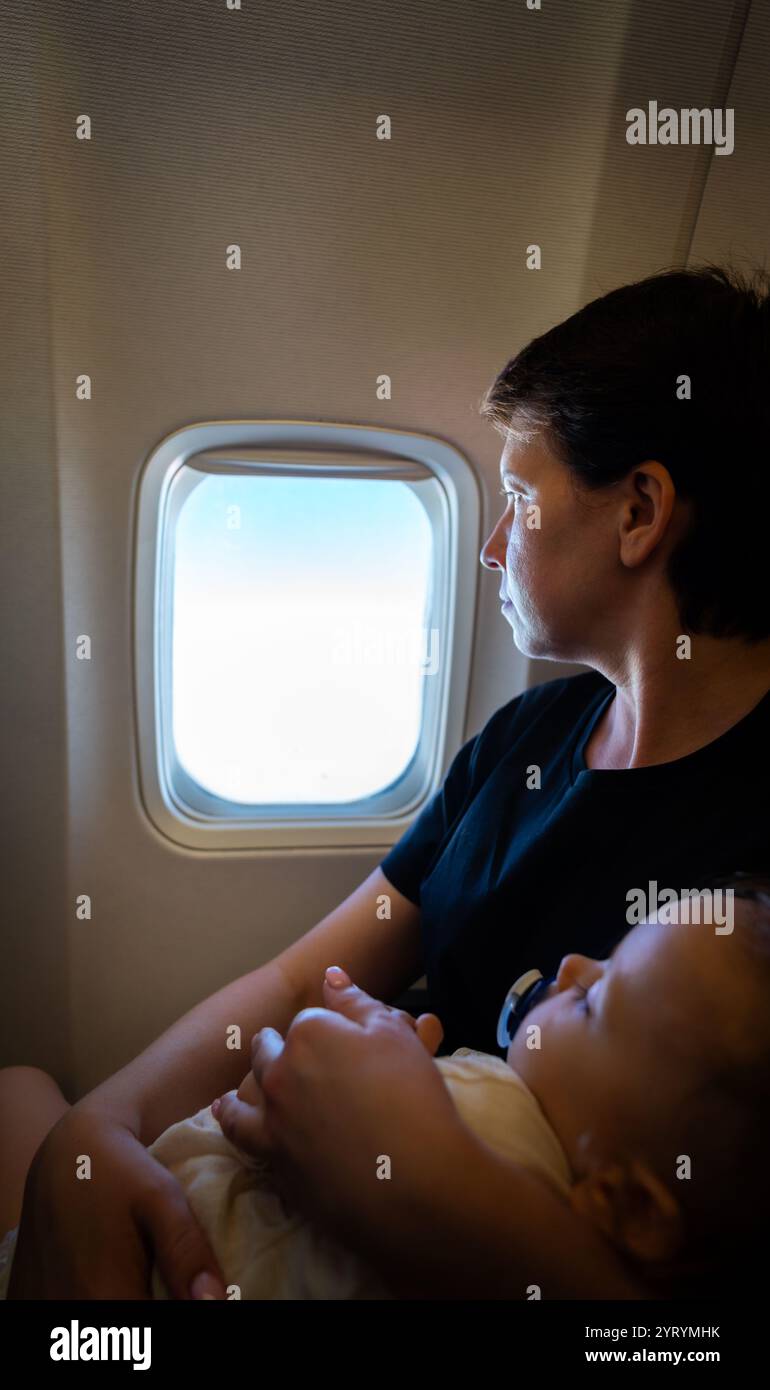 Mutter hält ein Kleinkind in der Hand, das im Flug aus dem Flugzeugfenster blickt. Konzept von Familienreisen und gemeinsamen Abenteuern Stockfoto