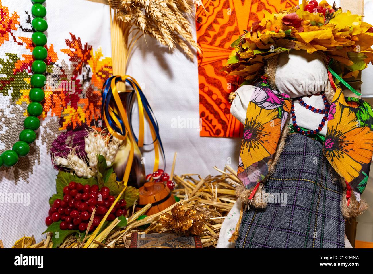 Eine handgefertigte Puppe mit leuchtenden Stoffen ist umgeben von Dekorationen mit Herbstmotiven, darunter Körner, Beeren und bunte Bänder, die eine Kuh einfangen Stockfoto