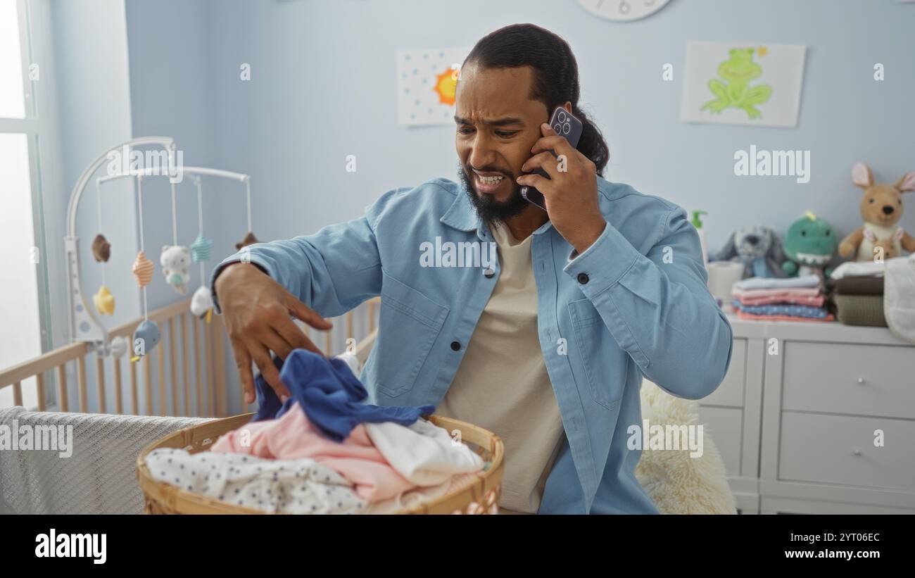 Mann, der telefoniert, während er Babykleidung in einem hellen Kinderzimmer mit Kinderbett und verspielter Einrichtung sortiert, bringt Fokus und Sorge drinnen zum Ausdruck. Stockfoto