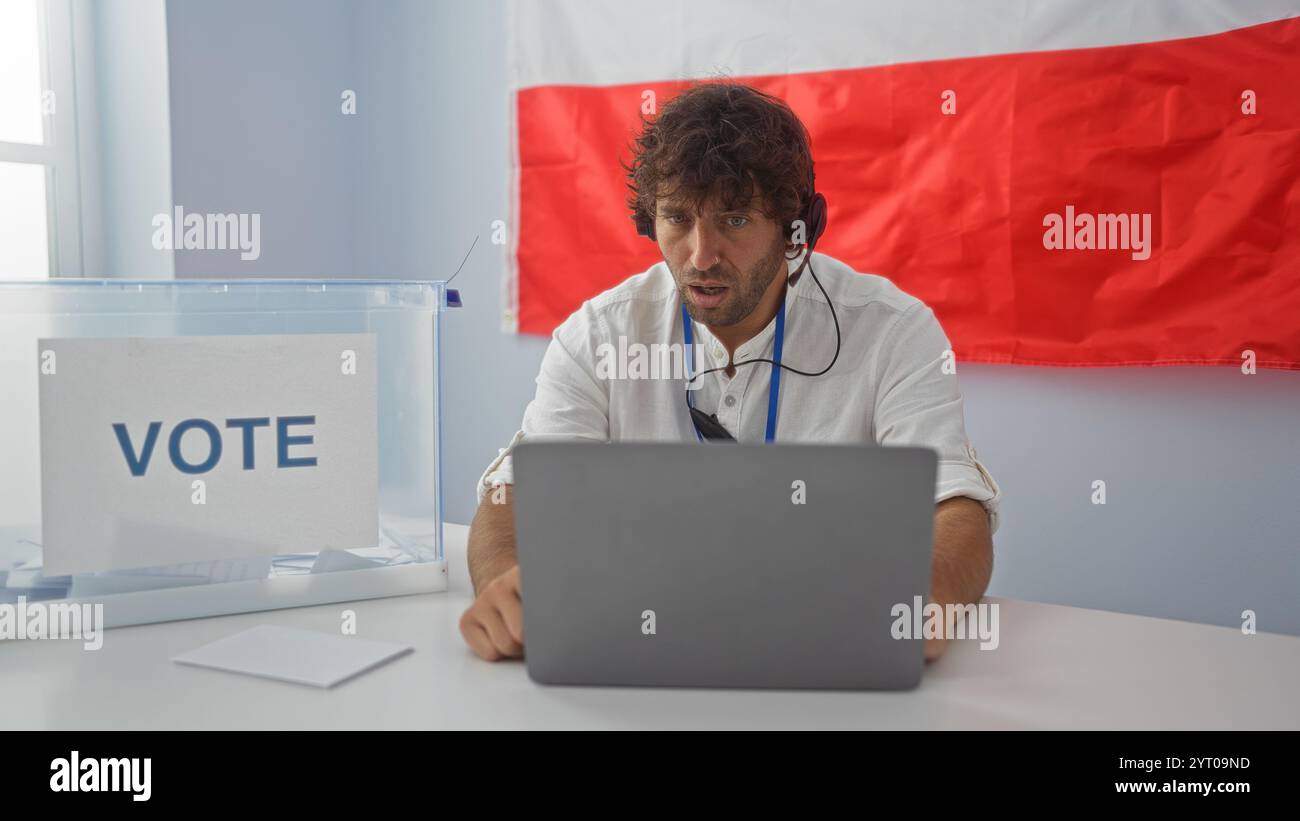 Junger Mann, der in einem Wahlzimmer mit einer polnischen Flagge im Hintergrund abstimmt, auf einen Laptop blickt und konzentriert erscheint. Stockfoto
