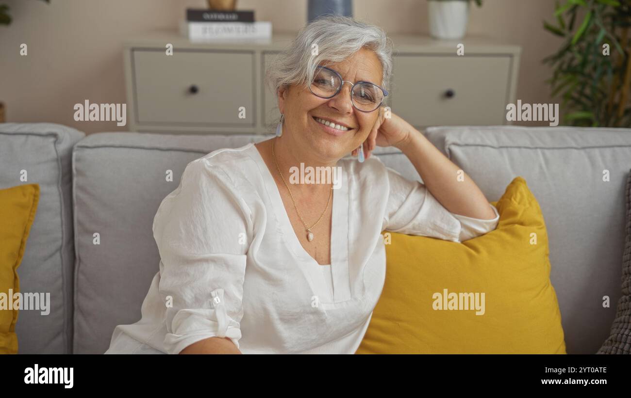 Grauhaarige Frau lächelt in einem gemütlichen Wohnzimmer mit gelben Kissen und zeigt eine schöne, reife Person mittleren Alters in einer Inneneinrichtung. Stockfoto