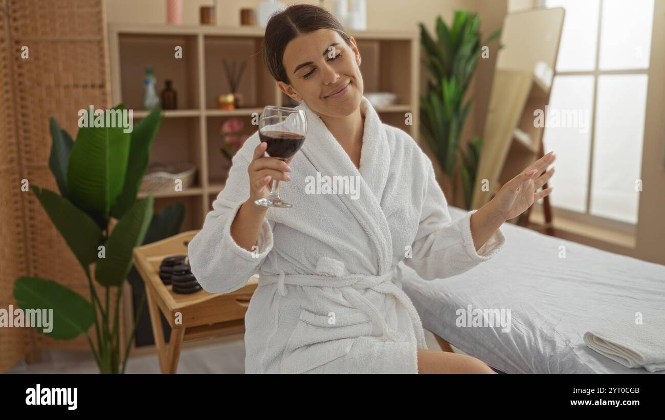 Junge Frau in einem Spa in einem weißen Bademantel, genießt ein Glas Wein, während sie lächelt und sich in einem ruhigen Wellness-Center mit Pflanzen und Dekorationen entspannt Stockfoto