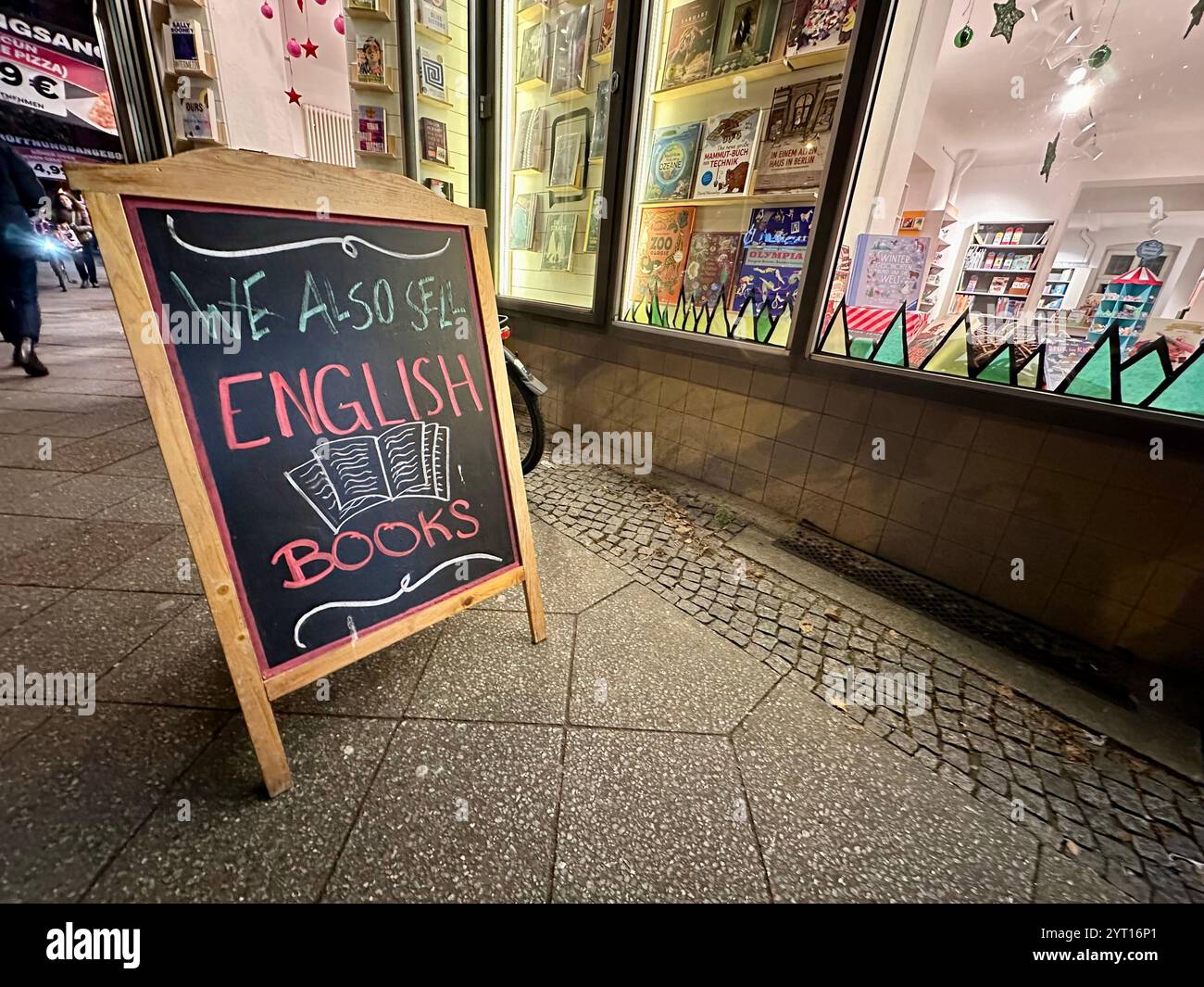 Eine hölzerne Tafel auf dem Bürgersteig Werbung 'Wir verkaufen auch englische Bücher' vor dem Buchladen Anagramm in Kreuzberg, Berlin Stockfoto