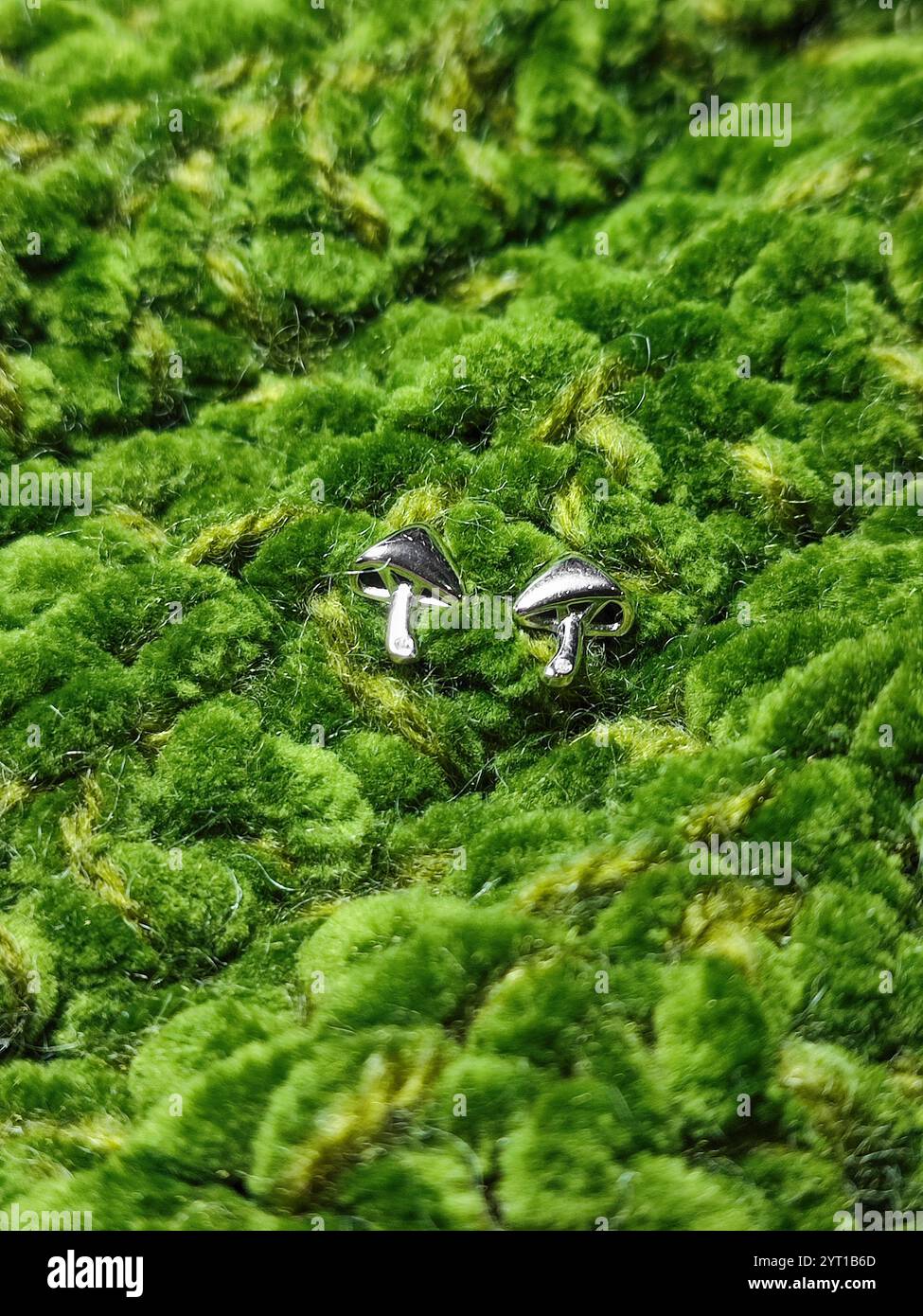 Kleine Pilz-Ohrringe auf grüner Strickoberfläche. Silberne Ohrringe für Damen. Pilze, pilzförmiger Schmuck. Schmuckset. Gestricktes Samtmuster Stockfoto