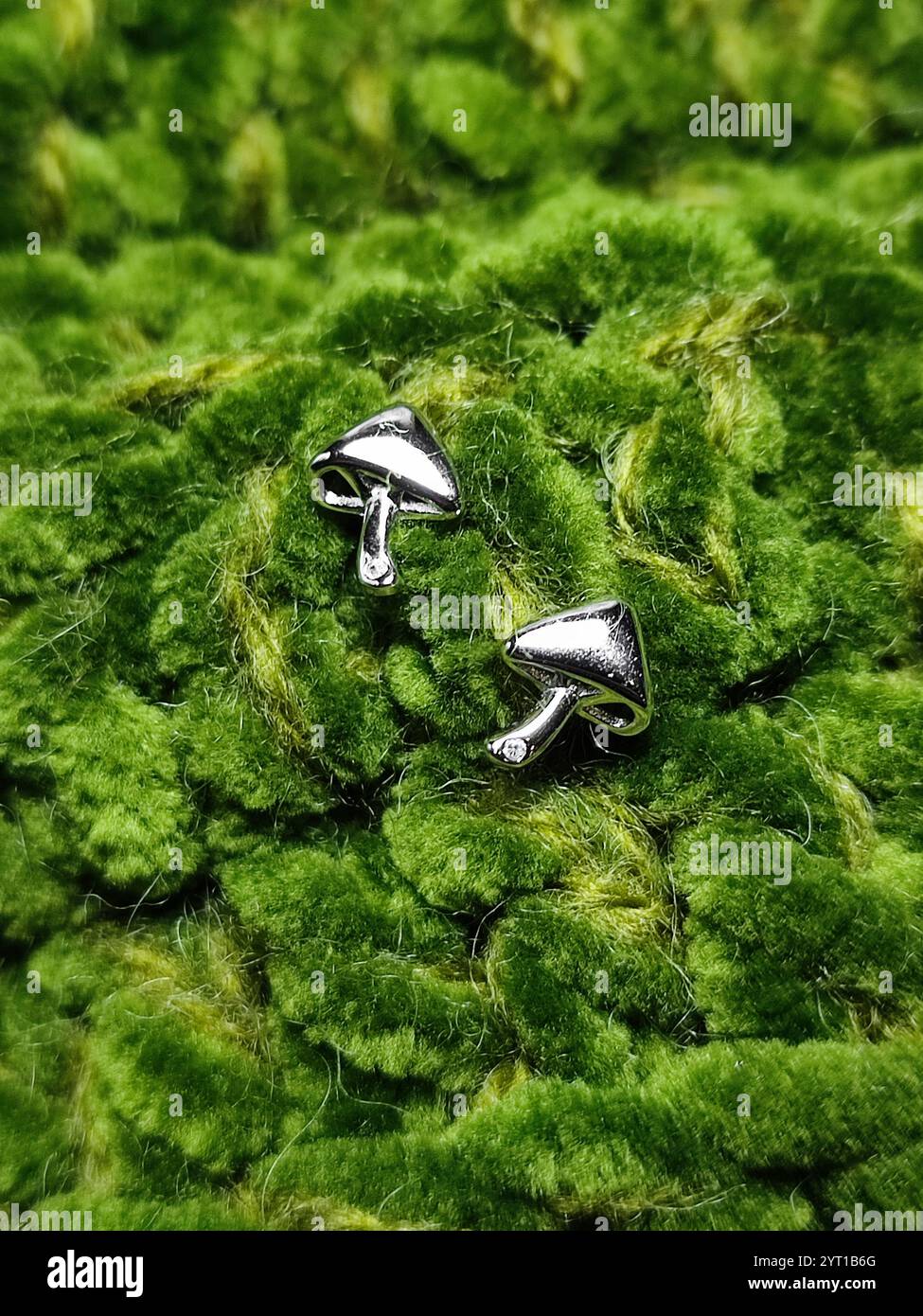 Kleine Pilz-Ohrringe auf grüner Strickoberfläche. Silberne Ohrringe für Damen. Pilze, pilzförmiger Schmuck. Schmuckset. Gestricktes Samtmuster Stockfoto