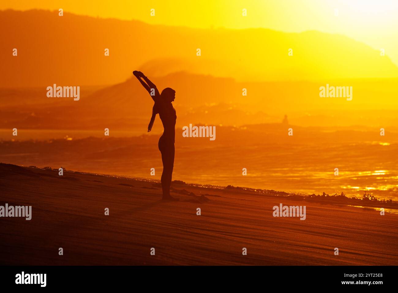 Dezember 2024. Ehukai Beach Park, Pupukea, Oahu, HI. Eine Frau dehnt sich mit Wellen und der untergehenden Sonne hinter ihr an der Nordküste von Oahu, HI. Stockfoto