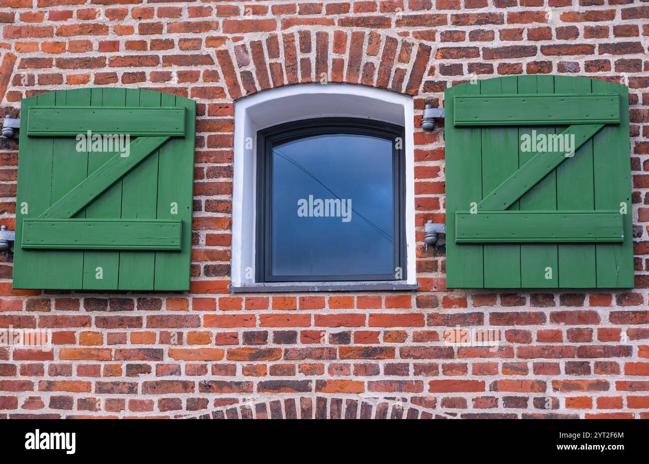 Eine malerische Ziegelmauer hat ein zentriertes Fenster mit einem weißen Rahmen, umgeben von grünen Holzläden Stockfoto