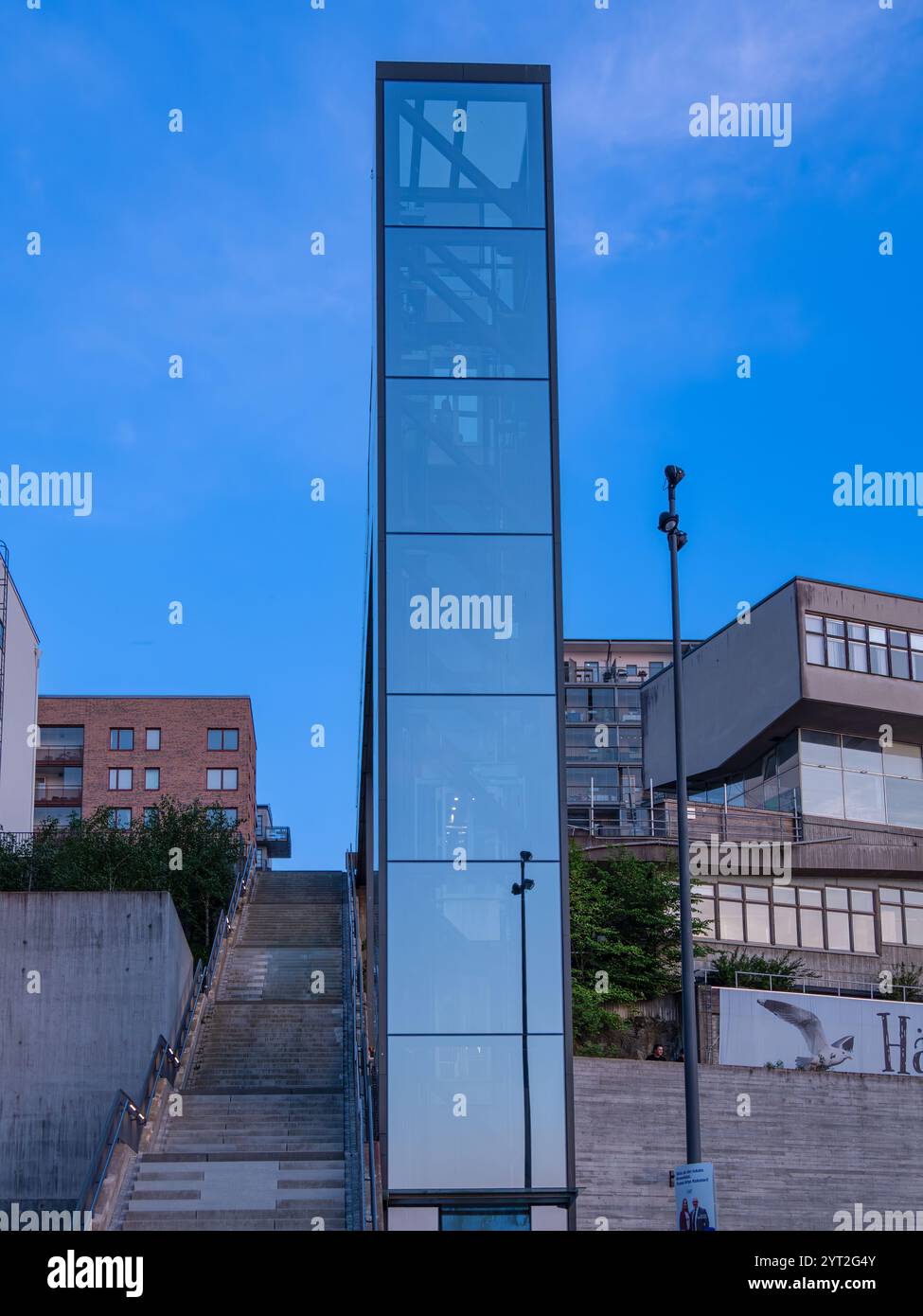 Stockholm, Schweden - 24. Mai 2024: Ein eleganter Glasaufzug verbindet die städtischen Ebenen, eingerahmt von moderner Architektur und Treppenhäusern. Stockfoto