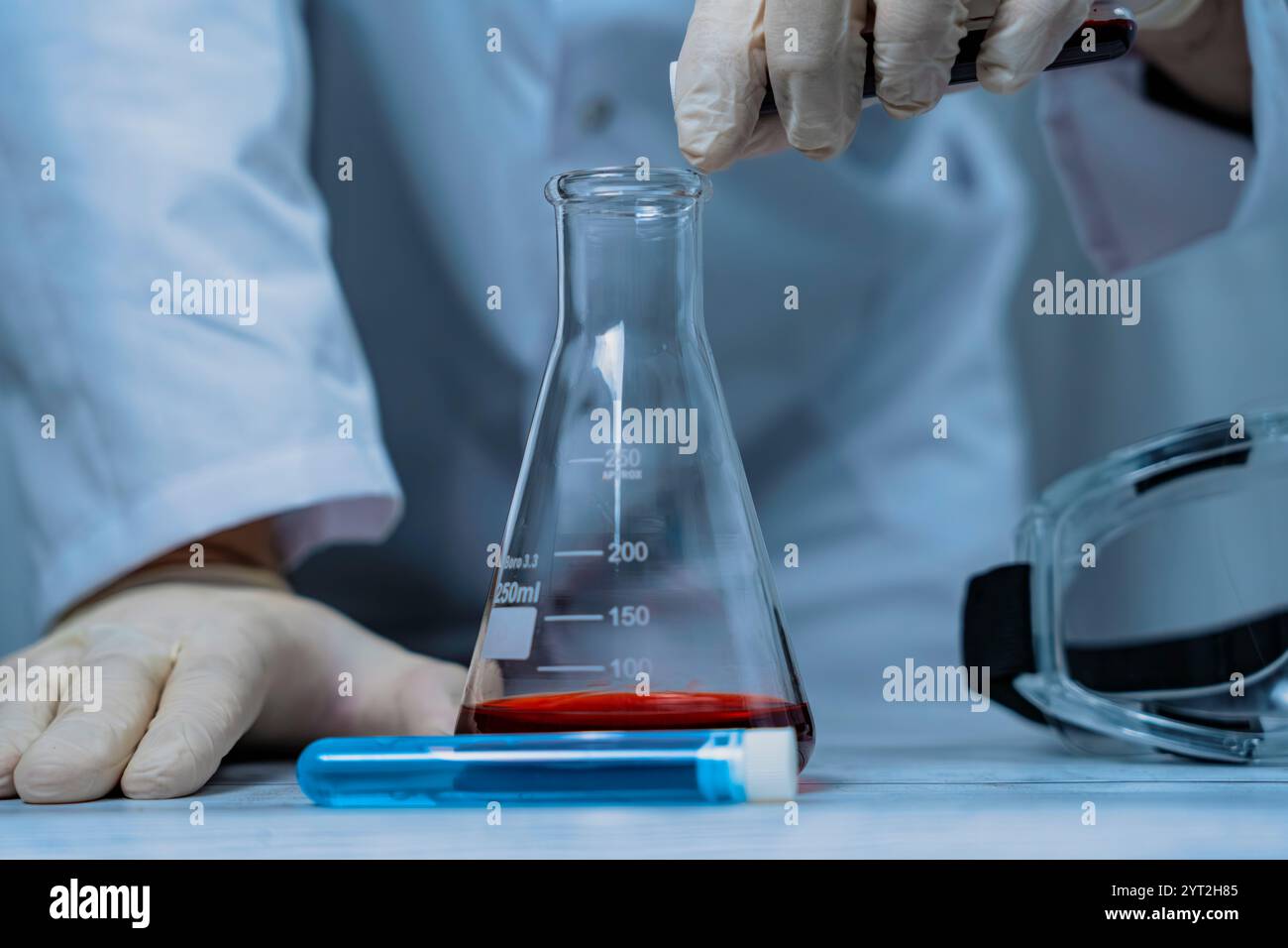 Ein Wissenschaftler hält ein mit grüner Flüssigkeit gefülltes Reagenzglas in einem Labor, umgeben von verschiedenen Laborgeräten. Das Bild hebt den Fokus und die Precis hervor Stockfoto