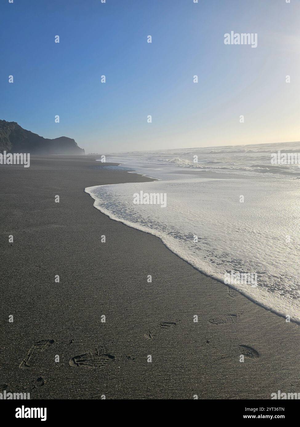 Ruhige Küstenlandschaft mit sanften Wellen, die über die Sandküste strömen, beleuchtet vom sanften Schein des Sonnenlichts unter einem klaren blauen Himmel. Stockfoto