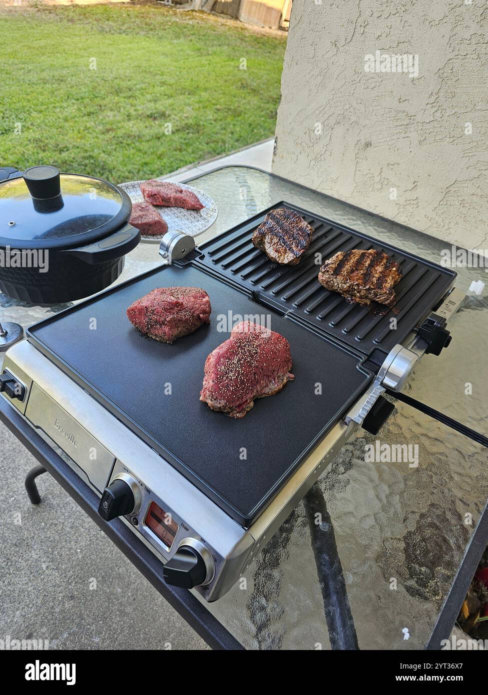 Grillszene im Freien mit rohen und gekochten Steaks auf einer Grillplatte, umgeben von einem grasbewachsenen Hinterhof und Terrassenbereich. Fängt einen entspannten Grillmoment ein. Stockfoto