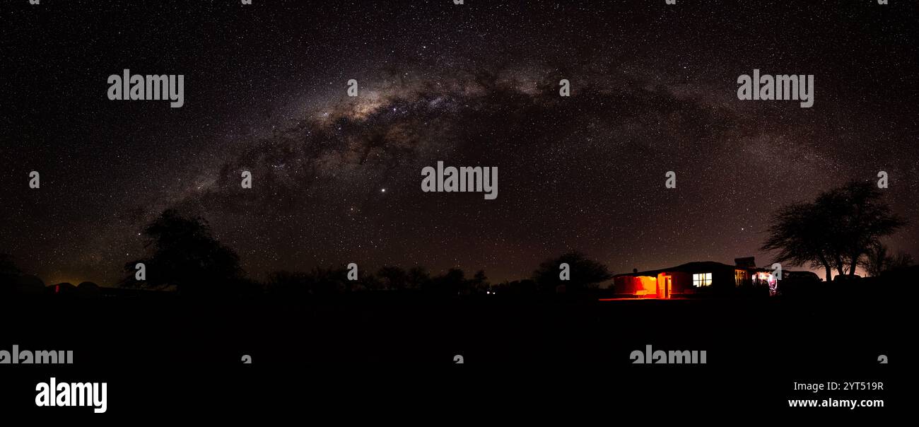 Atemberaubendes nächtliches Panorama von der Atacama-Wüste mit dem sternenübersäten Himmel und dem Bogen der Milchstraße. Ein kleines Haus und ein Baum auf dem Boden sorgen für Tiefe Stockfoto
