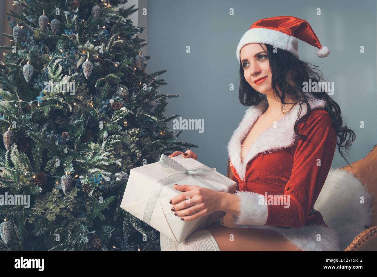 Ein Mädchen im Weihnachtskostüm träumt von einem Weihnachtsgeschenk. Eine junge Frau, die ein Geschenk auf dem Hintergrund eines Weihnachtsbaums hält und nachdenklich schaut Stockfoto