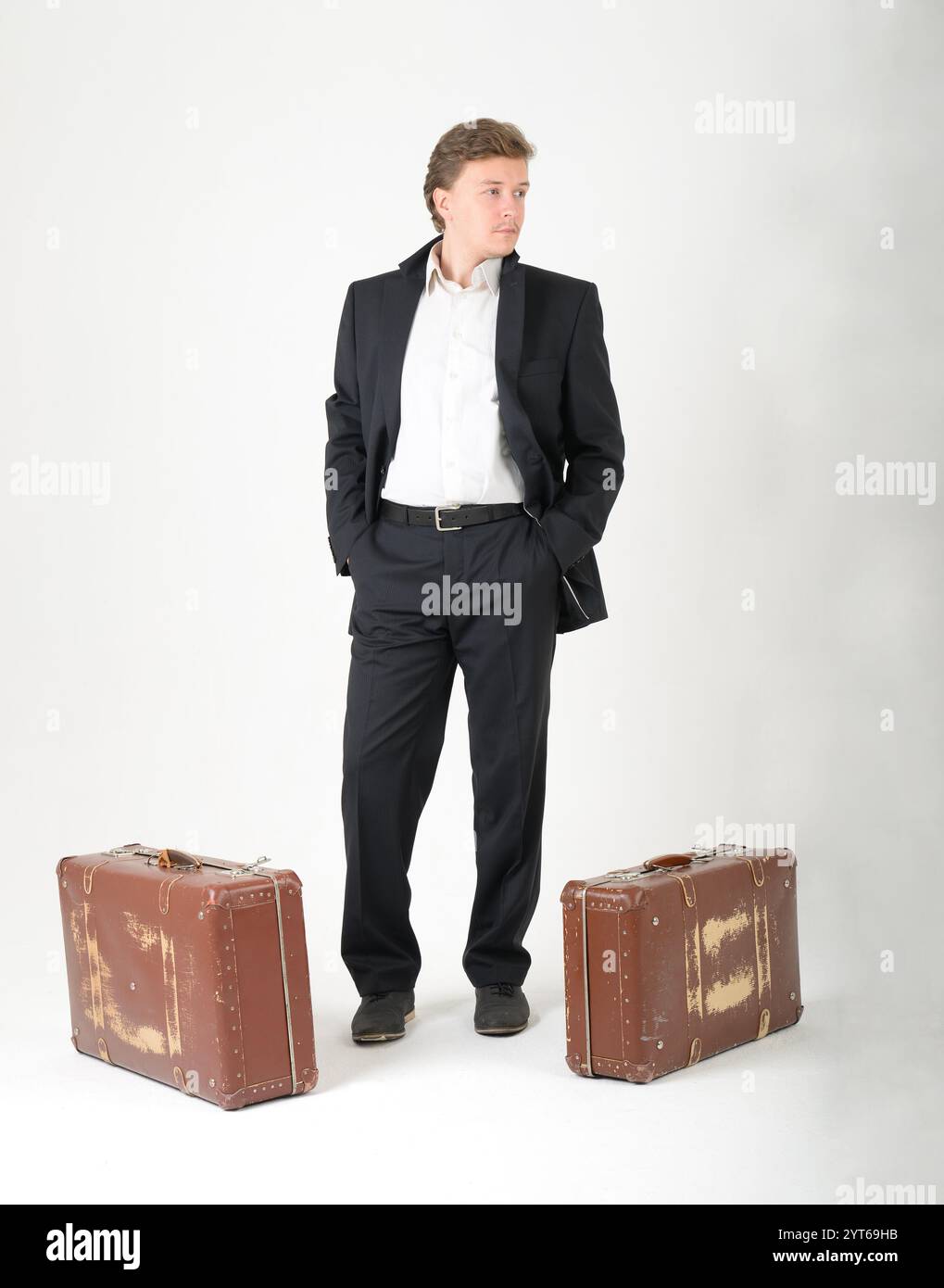 Ein langer Mann in schwarzem Anzug mit weißem Hemd und zwei Vintage-Koffern auf weißem Hintergrund. Das Konzept des Umzugs und der Verlagerung, das Konzept des Verzugs Stockfoto