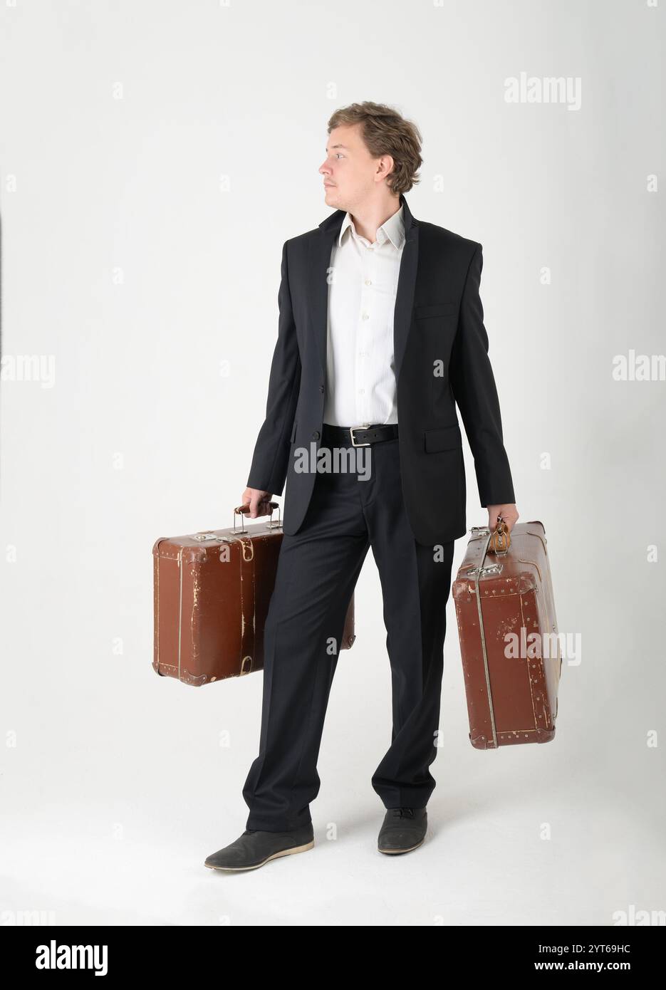 Langer Mann, der einen schwarzen Anzug mit einem weißen Hemd trägt und zwei Vintage-Koffer auf weißem Hintergrund hält. Das Konzept des Umzugs und der Verlagerung, das c Stockfoto