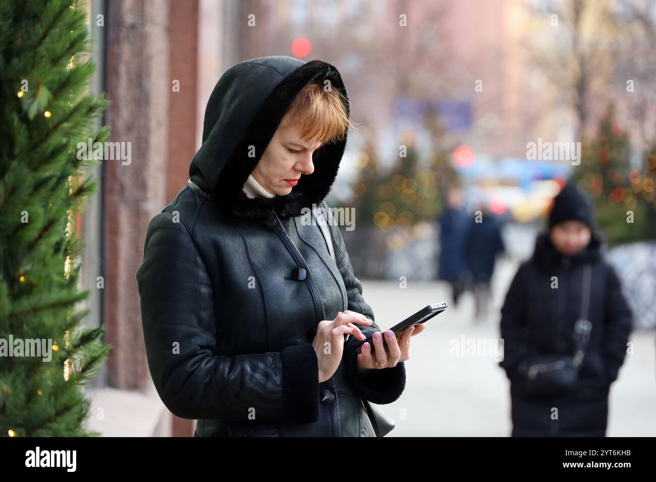 Frau mit warmem Mantel mit Smartphone auf der Stadtstraße auf dem Hintergrund von Neujahrsbäumen, mobile Kommunikation im Winter Stockfoto